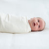 A baby in a HALO Swaddle Micro-Fleece TOG 3.0 rests on a plain surface, gazing upwards with wide eyes and an open mouth. Adjustable wings provide comfort and support healthy hip development, under soft lighting.
