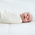 A baby in a HALO Swaddle Micro-Fleece TOG 3.0 rests on a plain surface, gazing upwards with wide eyes and an open mouth. Adjustable wings provide comfort and support healthy hip development, under soft lighting.