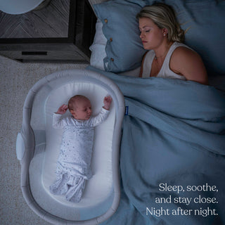 A woman sleeps in bed beside her baby, who rests comfortably in the HALO BassiNest® Soothing Swivel Sleeper 3.0 Bassinet. Both are under blue bedding. Text at the bottom reads, “Sleep, soothe, and stay close. Night after night.”.