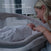 A woman smiles and touches her swaddled baby in the HALO BassiNest® Soothing Swivel Sleeper 3.0 Bassinet, featuring a mesh lowering wall. Text: Easy Lowering Wall—effortless access without bending or twisting for soothing comfort.