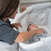 A woman gently places her hand on a sleeping baby in a gray-patterned BassiNest® Twin Sleeper by HALO. As the soothing vibrations ensure safe sleep, the woman watches joyfully indoors.