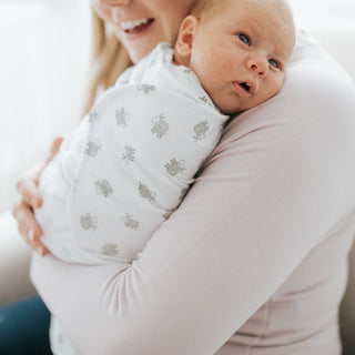 A smiling woman holds a newborn wrapped in a HALO Swaddle 100% Cotton TOG 1.5 with gray animal prints as the baby looks over her shoulder with wide, curious eyes.
