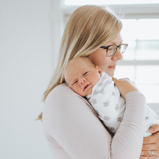 A woman with long blonde hair and glasses gently holds a newborn on her shoulder. The baby is wrapped in a HALO Swaddle 100% Cotton TOG 1.5 with a simple white pattern, as soft natural light fills the room.
