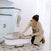 A woman kneels on a patterned tile floor in the laundry room, assembling the HALO BassiNest® Luxe Swivel Sleeper Bassinet beside the washer and dryer. She wears a beige cardigan and focuses on setting up the cozy bedside sleeper for her baby.