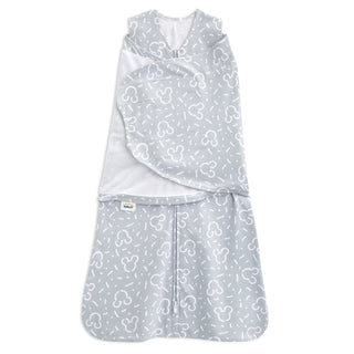 The HALO Disney™ 100% Cotton Swaddle TOG 1.5 is a light gray wearable blanket with white abstract shapes and Mickey Mouse outlines, a front zipper, and adjustable wings for cozy, safe sleep.