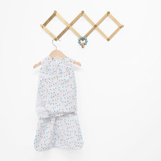 A HALO Swaddle Micro-Fleece TOG 3.0 with a colorful dot design hangs on a zigzag wooden wall rack, featuring blue balls on one hook. Its adjustable wings ensure comfort and healthy hip development, set against a plain white background.