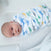 A baby lies peacefully in a HALO Swaddle Micro-Fleece TOG 3.0 with dinosaur patterns, gazing upwards. The adjustable wings ensure comfort, while the softly blurred background highlights the serene expression on the little ones face.