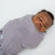 A baby is sleeping peacefully in a gray HALO 2-piece Organic Cotton Gift Box swaddle, snugly wrapped on a white background. The swaddle is made of organic cotton and features a visible tag.