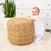 A baby in a white patterned 2-piece Organic Cotton Gift Box by HALO stands, leaning on a large woven ottoman. The setting includes a white dresser and tall green potted plant on a textured rug. The baby gazes upward, mouth slightly open.