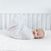 A baby lies on its back in a white crib, wrapped in a patterned white swaddle. The baby smiles while resting on the DreamWeave™ Breathable Crib Mattress by HALO, which features a soft, hypoallergenic quilted design.