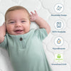 A smiling baby in a green outfit lies on the HALOs Breathable Nursery Bundle crib mattress, enjoying the quilted surface. Its features, like breathable design, 100% washability, hypoallergenic material, and GREENGUARD Gold certification, are highlighted with simple icons.