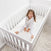 A young child wearing star-patterned white pajamas sits smiling in a white crib featuring the HALO DreamWeave™ Breathable Crib Mattress. The crib rests on a light carpet in a bright room with white walls. The child looks happy and playful.