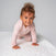 A young child with curly hair, in a light pink outfit, is crawling on the white textured surface of the HALO DreamWeave Breathable Crib Cover. The plain gray background accentuates their playful and curious expression as they explore their hypoallergenic world.
