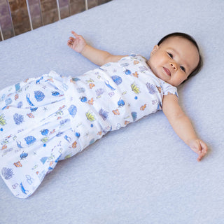 A baby lies contentedly on a light gray bed, wrapped in a HALO Finding Nemo Swaddle Cotton TOG 1.5. The white and blue-patterned sleeping bag features playful illustrations of leaves and animals, ensuring safe sleep. Beneath, the cozy light blue outfit complements the serene setting.