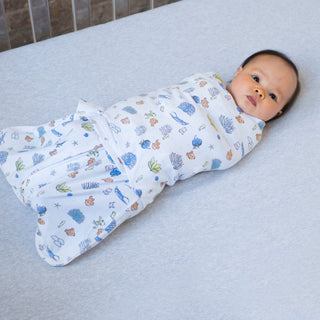 A baby lies on a gray surface, wrapped securely in a white HALO Finding Nemo Swaddle Cotton TOG 1.5 featuring cartoon animals and plants. The baby, with short dark hair and wide eyes, embodies safe sleep against a softly lit background, enhancing the peaceful scene.