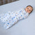 A baby lies on a gray surface, wrapped securely in a white HALO Finding Nemo Swaddle Cotton TOG 1.5 featuring cartoon animals and plants. The baby, with short dark hair and wide eyes, embodies safe sleep against a softly lit background, enhancing the peaceful scene.