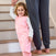 A cheerful young girl in a pink dinosaur-patterned outfit, similar to the HALO Toddler 100% Cotton TOG 0.5 design, smiles while holding hands with an adult in dark gray pants on wooden stairs, suggesting shes enjoying better sleep and cozy comfort.