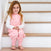 A young girl with long hair, in a pink and white outfit, sits smiling on wooden stairs. Her hands rest gently on her lap, bare feet touching the step below. The simple backdrop complements her tranquility—perhaps due to better sleep in her HALO Toddler 100% Cotton TOG 0.5 SleepSack.
