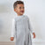A toddler stands indoors, happily looking up in a cozy light gray HALO Toddler Luxe Fleece TOG 2.0 with adjustable features over a long-sleeved white shirt, ensuring safer sleep against a plain light-colored background.