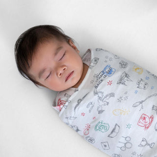 A dark-haired baby is peacefully resting on a soft, white surface, wrapped in the whimsical Harry Potter™ Swaddle by HALO, made of 100% cotton with a TOG rating of 1.5.