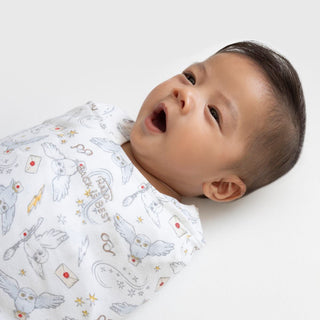 A baby lies on a white surface, wrapped in the HALO Harry Potter™ Swaddle 100% Cotton TOG 1.5. The swaddle features owl, envelope, and glasses designs with adjustable Velcro fasteners to support healthy hip development as the little one yawns with eyes wide open.