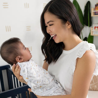 A smiling woman holds a baby in a Harry Potter™ Swaddle 100% Cotton TOG 1.5 by HALO. She wears a white sleeveless top in a room with a crib and books, as adjustable Velcro fasteners promote healthy hip development.