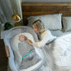 A woman lies in bed, leaning on her side, looking at a baby wrapped in a HALO Swaddle Ideal Temp TOG 1.5 with a 3-way swaddle design inside a bedside bassinet. The room features a warm lamp on the nightstand and a wooden headboard.