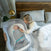 A woman lies in bed, leaning on her side, looking at a baby wrapped in a HALO Swaddle Ideal Temp TOG 1.5 with a 3-way swaddle design inside a bedside bassinet. The room features a warm lamp on the nightstand and a wooden headboard.