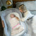 A woman rests on a bed beside a baby in a cozy bassinet, snugly wrapped in the HALO Swaddle Ideal Temp TOG 1.5 with pink accents. A lamp and small plant on the bedside table enhance the serene ambiance.
