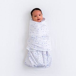 A baby is comfortably wrapped in a HALO Swaddle 100% Cotton TOG 1.5 with adjustable wings, featuring a white blanket with blue stars. Resting on a light surface, the babys face is visible, looking upward while promoting healthy hip development.