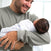 A man in a light green sweater tenderly holds a sleeping baby wrapped in a HALO Swaddle 100% Cotton TOG 1.5 with blue stars, designed for healthy hip development. He smiles warmly at the baby while standing in a softly lit room.