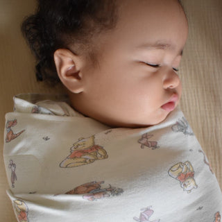A sleeping baby is snugly wrapped in a HALO Disney™ Winnie the Pooh 100% Cotton Swaddle TOG 1.5, resting on a soft surface with eyes closed and a serene expression.