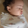 A sleeping baby is snugly wrapped in a HALO Disney™ Winnie the Pooh 100% Cotton Swaddle TOG 1.5, resting on a soft surface with eyes closed and a serene expression.
