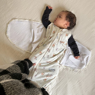 A baby rests on a light-colored sheet in a Disney™ Winnie the Pooh 100% Cotton Swaddle TOG 1.5 by HALO. Wearing a black long-sleeve shirt, the baby looks to the side as an adult gently adjusts the cozy swaddle.