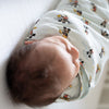A newborn peacefully sleeps on a soft white surface, wrapped snugly in the HALO Disney™ 100% Cotton Swaddle TOG 1.5, featuring Mickey and Minnie prints, viewed from an elevated angle.