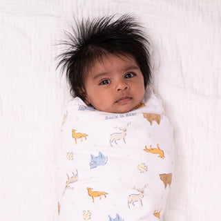 A dark-haired baby is cozily wrapped in a HALO Swaddle 100% Cotton TOG 1.5 with animal illustrations like deer and foxes, lying on a white surface. The adjustable wings ensure comfort and support healthy hip development while the baby gazes calmly at the camera.