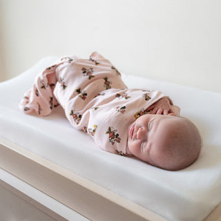 A baby peacefully sleeps on a changing table, wrapped in a light pink HALO Disney™ 100% Cotton Swaddle TOG 1.5, adorned with Mickey and Minnie. Resting safely on its back with one hand near its face, the serene scene unfolds against a plain light-colored wall.