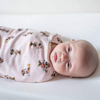 A baby lies on a white surface wrapped in a Disney™ 100% Cotton Swaddle TOG 1.5, featuring Mickey and Minnie. Their eyes are open with a calm expression, enjoying the cozy comfort synonymous with HALOs swaddles.