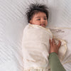 A baby with dark hair is wrapped in a cream-colored HALO Swaddle 100% Cotton TOG 1.5, lying on a textured white surface. An adult hand gently adjusts its innovative wings, ensuring the baby looks content and relaxed while promoting healthy hip development.