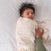 A baby with dark hair is wrapped in a cream-colored HALO Swaddle 100% Cotton TOG 1.5, lying on a textured white surface. An adult hand gently adjusts its innovative wings, ensuring the baby looks content and relaxed while promoting healthy hip development.