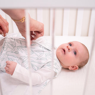 A baby sleeps in a HALO Swaddle 100% Cotton TOG 1.5 featuring a leaf pattern. Gentle hands adjust the swaddles wings for comfort and hip health, as white crib bars frame this tender scene.