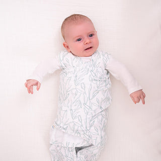 A baby in a HALO Swaddle 100% Cotton TOG 1.5 with adjustable wings lies on a soft white surface, looking upwards. The baby, with short hair and a long-sleeved white shirt under the swaddle, enjoys healthy hip development support.