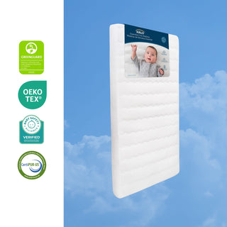 A rectangular, breathable HALO Essential Breathable Crib Mattress standing upright against a blue sky, with four GREENGUARD Gold certified safety logos to the left.
