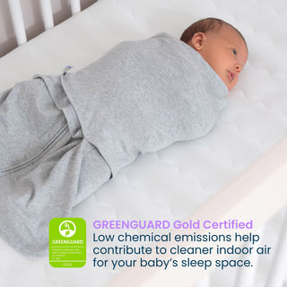 A baby swaddled in gray fabric lies on a HALO Essential Breathable Crib Mattress, with a graphic stating: Low chemical emissions help contribute to cleaner indoor air for your baby’s sleep space.