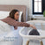 A woman in bed reaches for a baby in the HALO BassiNest® Essential Sleeper with mesh walls, as sunlight streams through the window. Text says, Soothe them from bed.