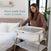 A person stands by a bed, adjusting the HALO BassiNest® Essential Sleeper with wooden legs and white mesh walls. The room features a large window, plant, and light-colored walls. Text on the image reads, Easily move this portable sleep space from room to room.