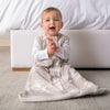 A baby sits on a gray carpet in front of a bed frame, wearing the HALO Wearable Blanket Ideal Temp TOG 1.0 over a white long-sleeve shirt. Utilizing IntelliThread technology for optimal temperature control, the baby smiles and claps hands with a cheerful expression.
.