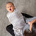 A baby wrapped in a HALO Swaddle Ideal Temp TOG 1.5 is yawning and looking upwards on a womans lap. She sits comfortably on the gray carpeted floor, wearing a gray sweater and black pants.