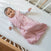 A baby lies in a crib on a white fitted sheet, wearing the aden + anais Bamboo Baby Bundle over a white long-sleeve shirt, looking up with one arm slightly raised.