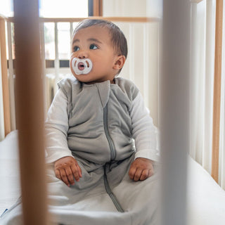 A toddler in a crib enjoys warmth wrapped in HALOs Wearable Blanket SuperSoft Bamboo TOG 1.5, complete with a convenient 2-way zipper. Sunlight streams through the window, adding to the calm and cozy ambiance.